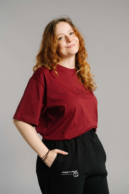 Burgunderrotes T-Shirt mit tonaler Stickerei des „T“-Logos im Lorbeerkranz auf der Brust. Das Shirt hat einen lockeren, gerade fallenden Schnitt mit Rundhalsausschnitt und leicht überschnittenen Schultern. Es liegt locker über einer schwarzen Jogginghose mit weißem Schriftzug auf dem Oberschenkel.