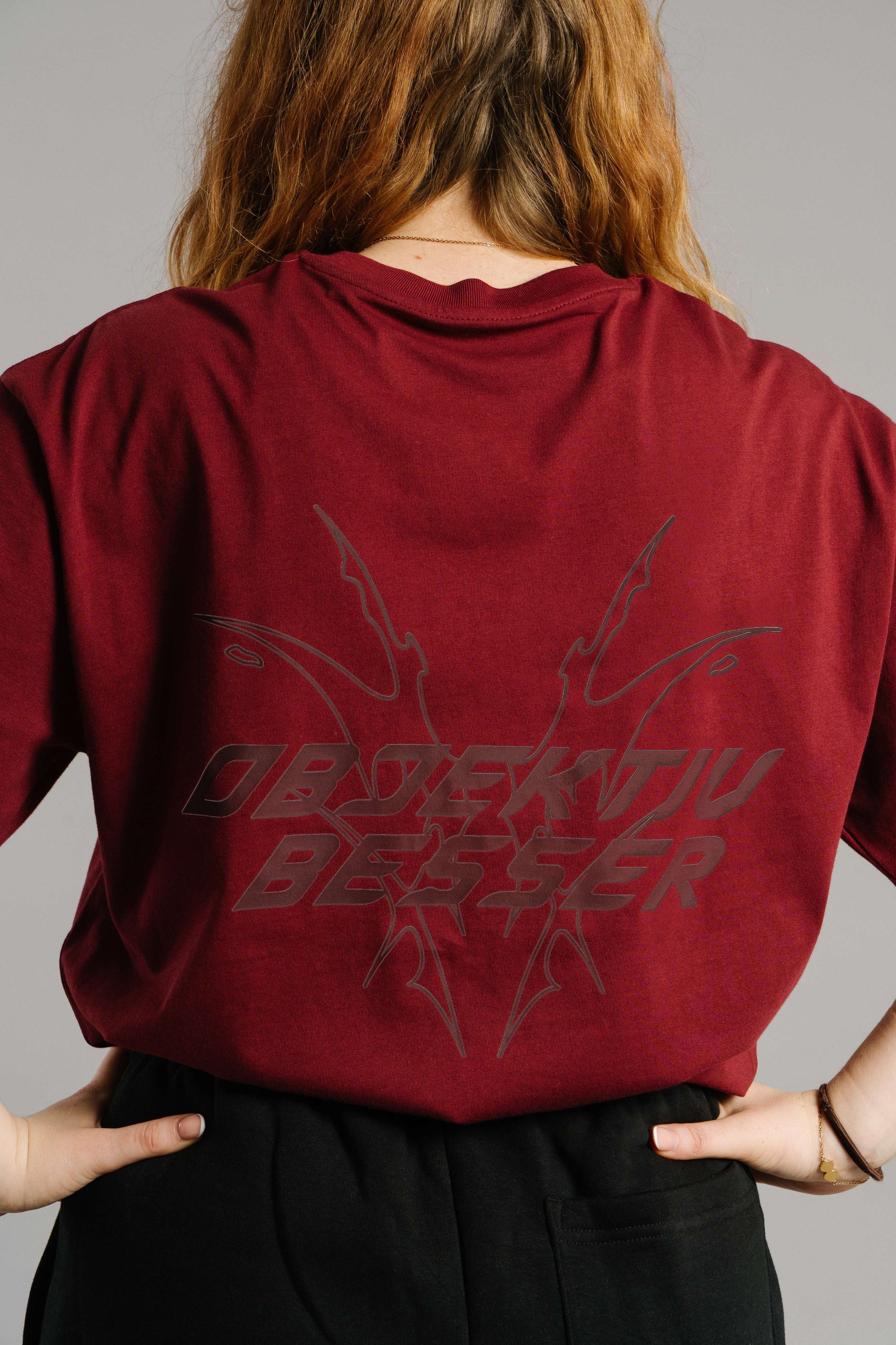 Burgunderrotes T-Shirt mit großem Rückendruck in dunkler Schrift. Das Motiv zeigt den typografischen Schriftzug „OBJEKTIV BESSER“ in markanter, futuristischer Schrift mit spitzen, grafischen Linienformen, die sich nach außen verzweigen. Der Stoff liegt locker über einer schwarzen Hose, die Hände sind lässig in die Gesäßtaschen gelegt.