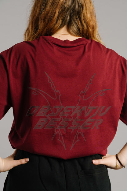 Burgunderrotes T-Shirt mit großem Rückendruck in dunkler Schrift. Das Motiv zeigt den typografischen Schriftzug „OBJEKTIV BESSER“ in markanter, futuristischer Schrift mit spitzen, grafischen Linienformen, die sich nach außen verzweigen. Der Stoff liegt locker über einer schwarzen Hose, die Hände sind lässig in die Gesäßtaschen gelegt.