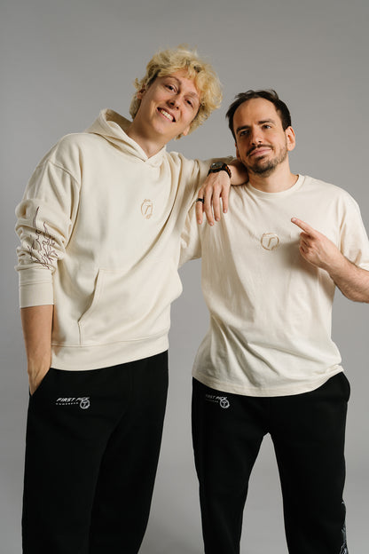 Zwei Personen stehen nebeneinander und lachen. Die Person rechts trägt ein beiges T-Shirt mit einem dezent eingestickten Logo auf der Brust – ein rundes Emblem mit dem Buchstaben „T“ und kleinen Lorbeerblättern. Sie zeigt mit dem Finger auf das Logo, wodurch der Fokus auf das gestickte Detail gelenkt wird.