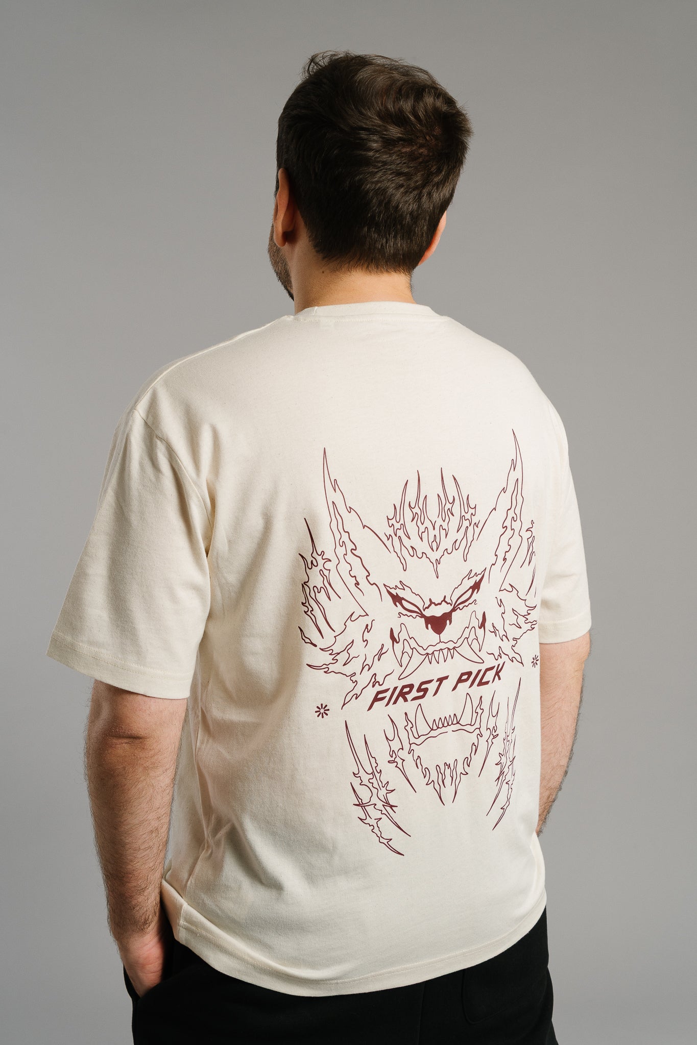 Eine Person trägt ein beiges T-Shirt mit großem Rückendruck in dunkelroter Liniengrafik. Das Motiv zeigt ein fauchendes, flammenförmiges Monstergesicht mit spitzen Ohren und Zähnen. Unterhalb der Illustration steht der Schriftzug „FIRST PICK“. Das Design wirkt dynamisch und kontrastiert auffällig mit der hellen Stofffarbe.