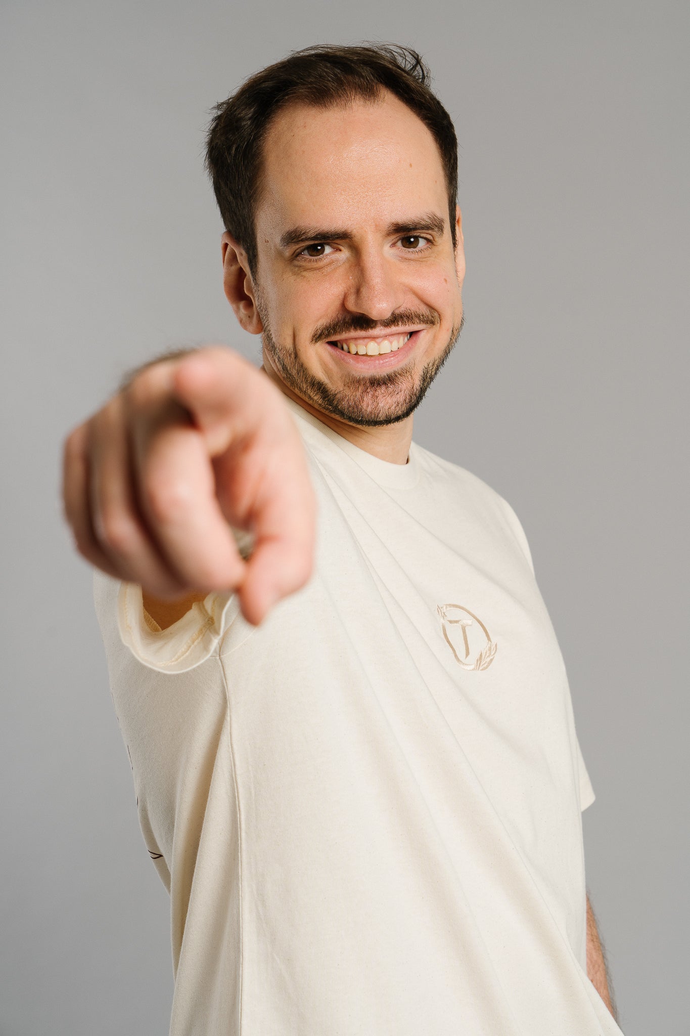 Eine Person trägt ein beiges T-Shirt mit kleinem gesticktem Logo auf der Brust – ein rundes Emblem mit dem Buchstaben „T“ und Lorbeerblättern. Die Person zeigt lächelnd und direkt in die Kamera, wodurch das Motiv einen humorvollen, selbstbewussten Ausdruck bekommt. Der Fokus liegt auf dem minimalistischen Logo und der lockeren Ausstrahlung.