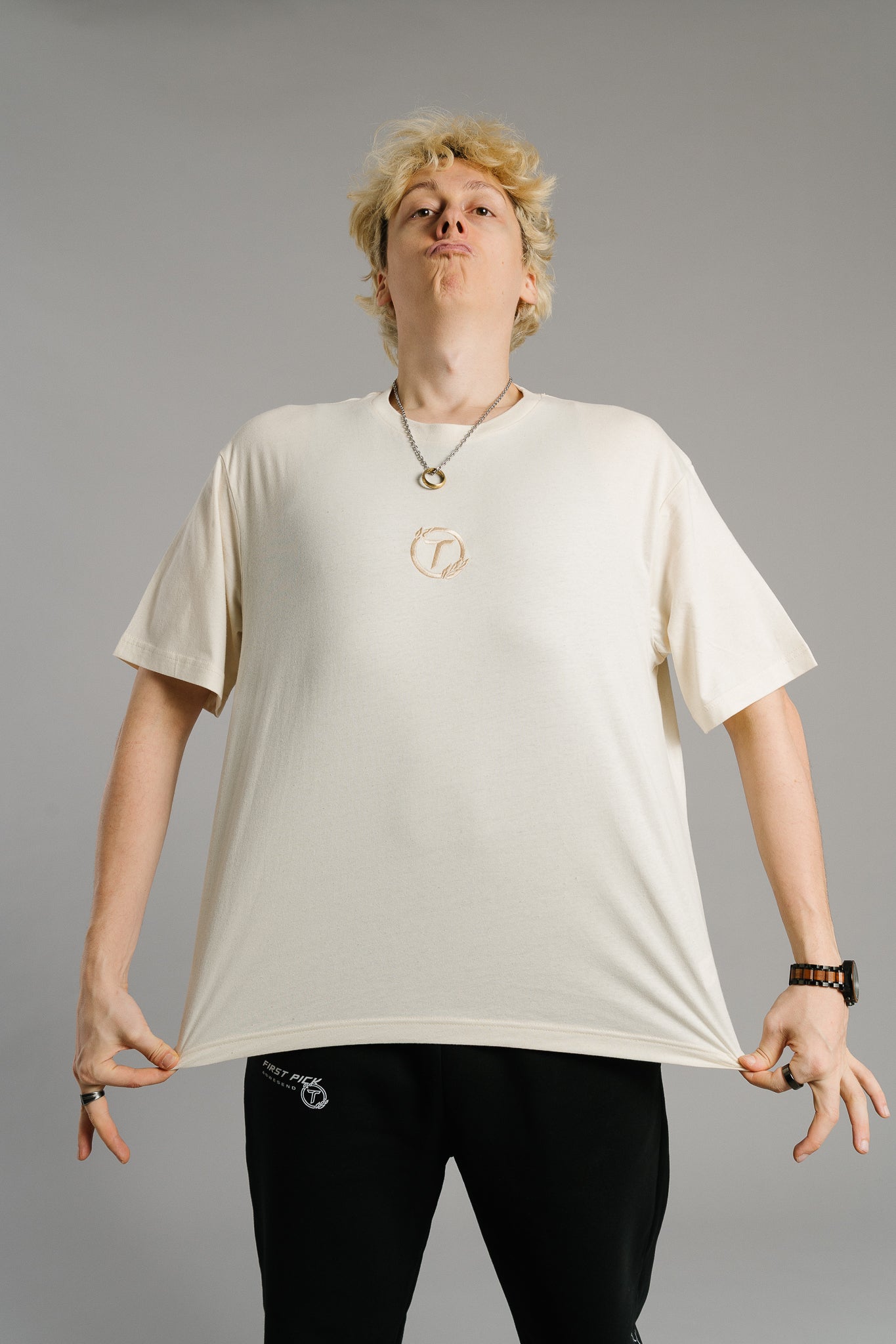 Eine Person trägt ein beiges T-Shirt mit kleinem gesticktem Logo auf der Brust – ein runder Lorbeerkranz mit dem Buchstaben „T“ in der Mitte. Das Shirt hat einen lockeren Schnitt und fällt weich. Die Person steht frontal zur Kamera, zieht den Stoff an beiden Seiten leicht nach außen und streckt das Shirt, um dessen Passform zu zeigen. Der Gesichtsausdruck wirkt selbstbewusst und spielerisch.
