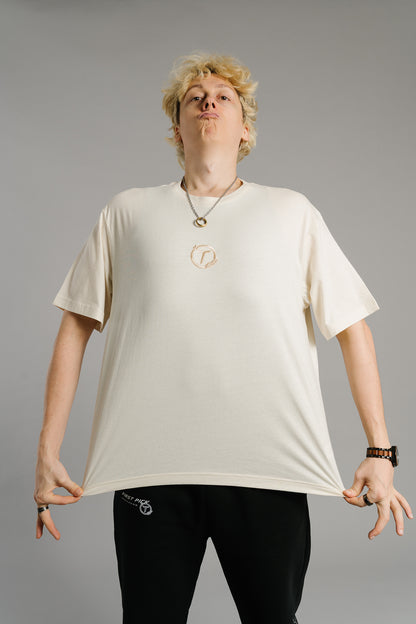 Eine Person trägt ein beiges T-Shirt mit kleinem gesticktem Logo auf der Brust – ein runder Lorbeerkranz mit dem Buchstaben „T“ in der Mitte. Das Shirt hat einen lockeren Schnitt und fällt weich. Die Person steht frontal zur Kamera, zieht den Stoff an beiden Seiten leicht nach außen und streckt das Shirt, um dessen Passform zu zeigen. Der Gesichtsausdruck wirkt selbstbewusst und spielerisch.
