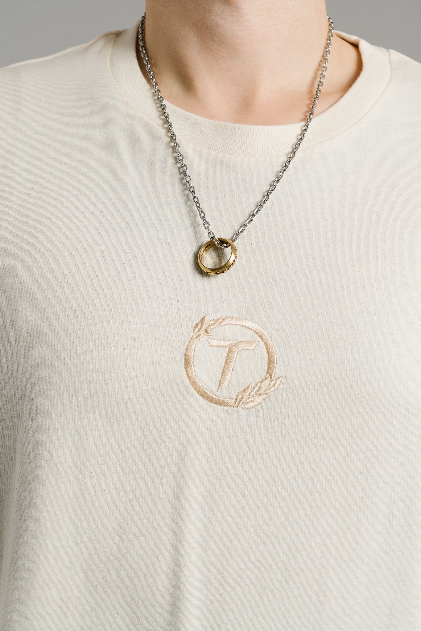 Nahaufnahme eines beigen T-Shirts mit gesticktem Brustlogo. Das Logo zeigt den Buchstaben „T“ in einem Kreis, der von einem Lorbeerzweig umrahmt wird, Ton in Ton in hellbeiger Stickerei. Der Stoff wirkt weich und leicht strukturiert. Im oberen Bildbereich ist eine silberne Kette mit einem goldfarbenen Ringanhänger zu sehen. Der Fokus liegt auf der Stickerei des Shirts.