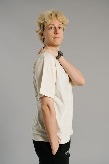 Eine Person trägt ein beiges T-Shirt mit lockerem Schnitt und kurzem Ärmel. Auf der Brust ist dezent das gestickte Logo mit dem Buchstaben „T“ im Lorbeerkranz zu sehen. Der Stoff wirkt weich und natürlich. Die Person steht seitlich zur Kamera, legt eine Hand entspannt auf die gegenüberliegende Schulter und blickt ruhig nach vorn.