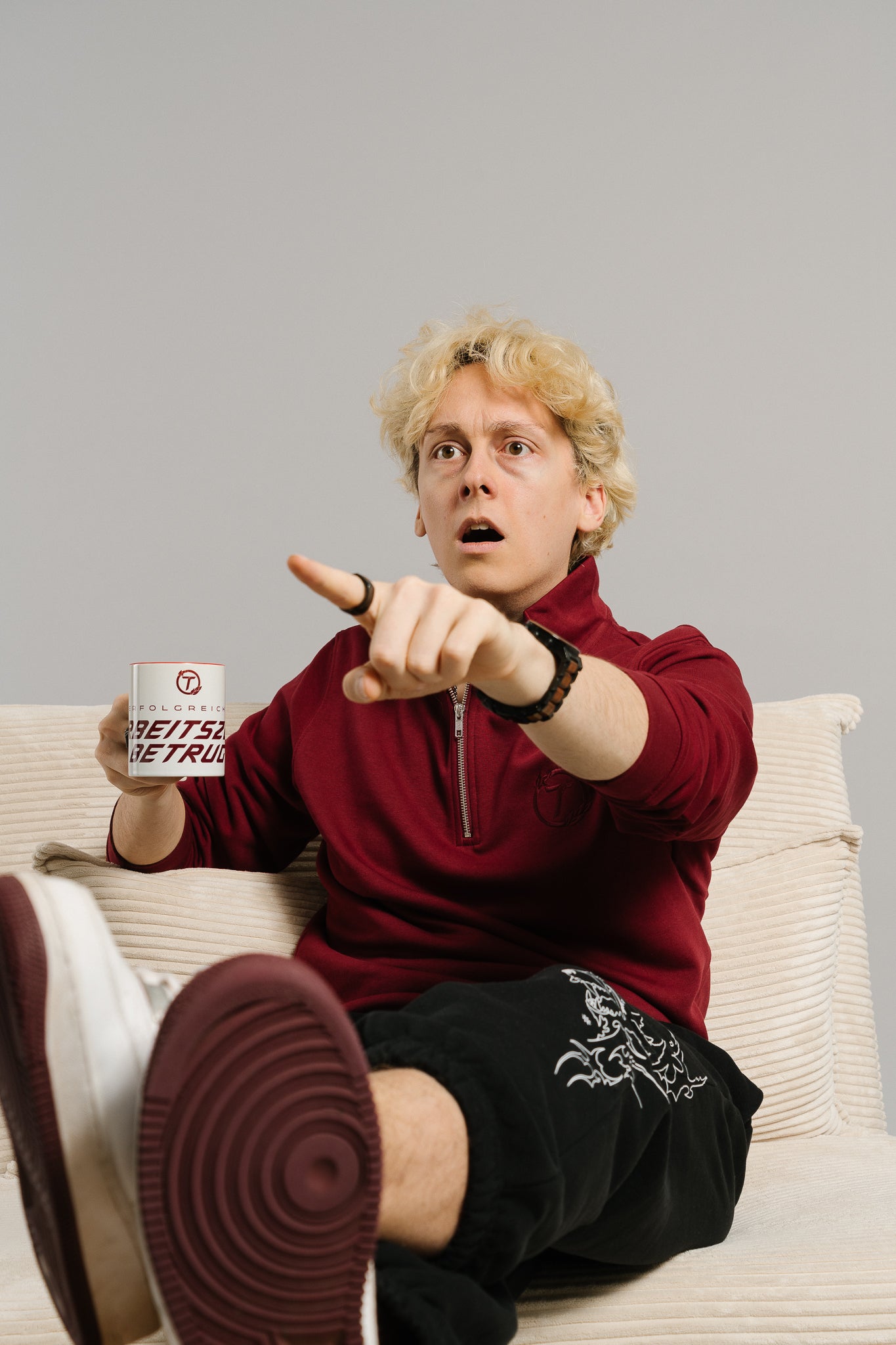 Eine Person mit blondem Haar sitzt lässig auf einem cremefarbenen Sofa und hält eine weiße Tasse mit rotem Innenbereich und dem sichtbaren Schriftzug „ERFOLGREICHER ARBEITSZEITBETRUG“ in der linken Hand. Mit erstaunter Miene zeigt sie mit der rechten Hand nach vorne, als hätte sie gerade etwas Unerwartetes entdeckt. Der humorvolle Ausdruck und die Pose verleihen dem Bild eine spontane, lebendige Wirkung, während die Tasse klar im Mittelpunkt steht.