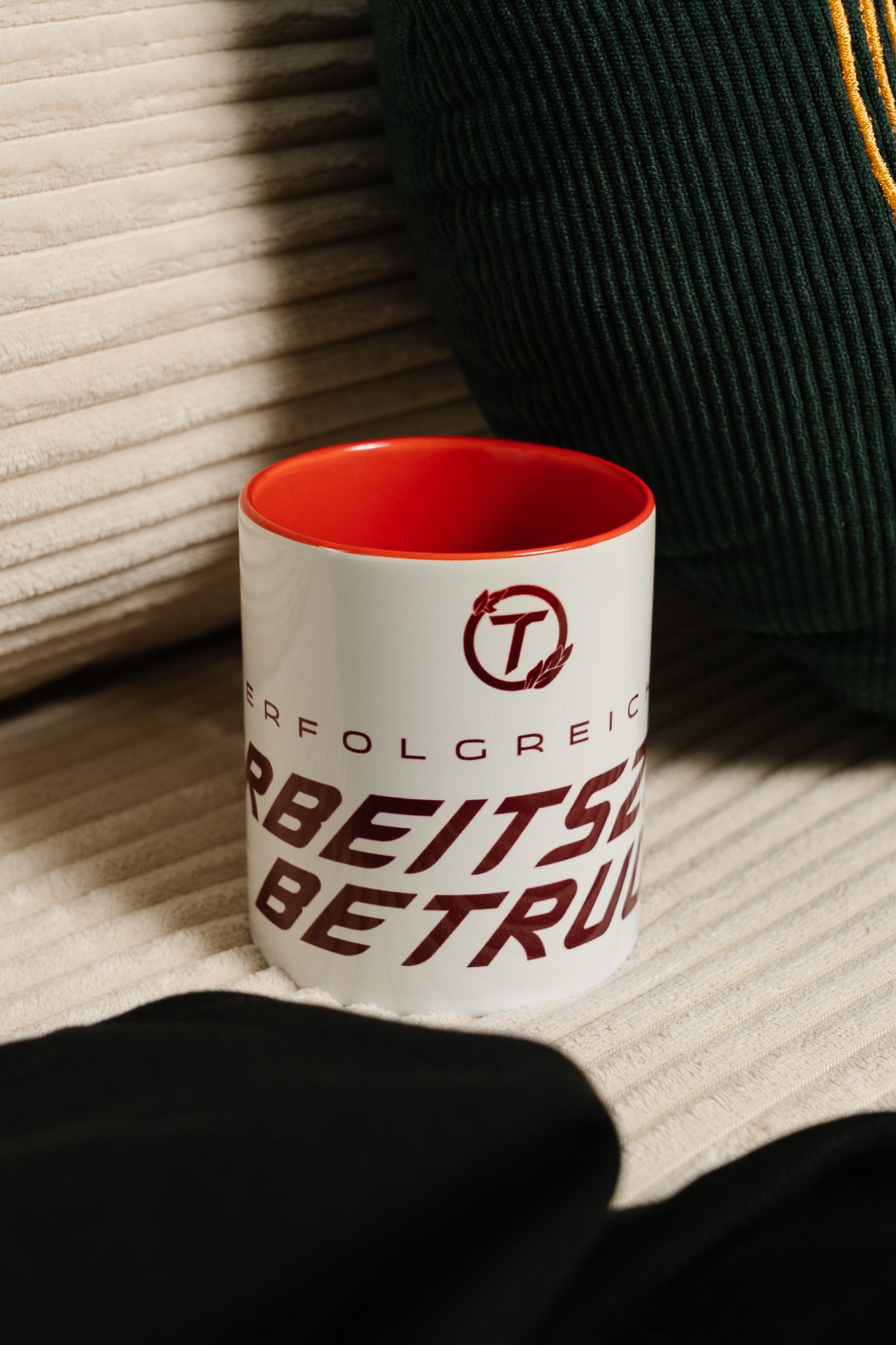Weiße Keramiktasse mit rotem Innenbereich und rotem Schriftzug „ERFOLGREICHER ARBEITSZEITBETRUG“. Die Tasse steht auf einem beigefarbenen, gerippten Sofa neben einem dunkelgrünen Kissen mit gelber Ziernaht. Durch das warme Licht entsteht eine gemütliche, leicht humorvolle Alltagsszene.