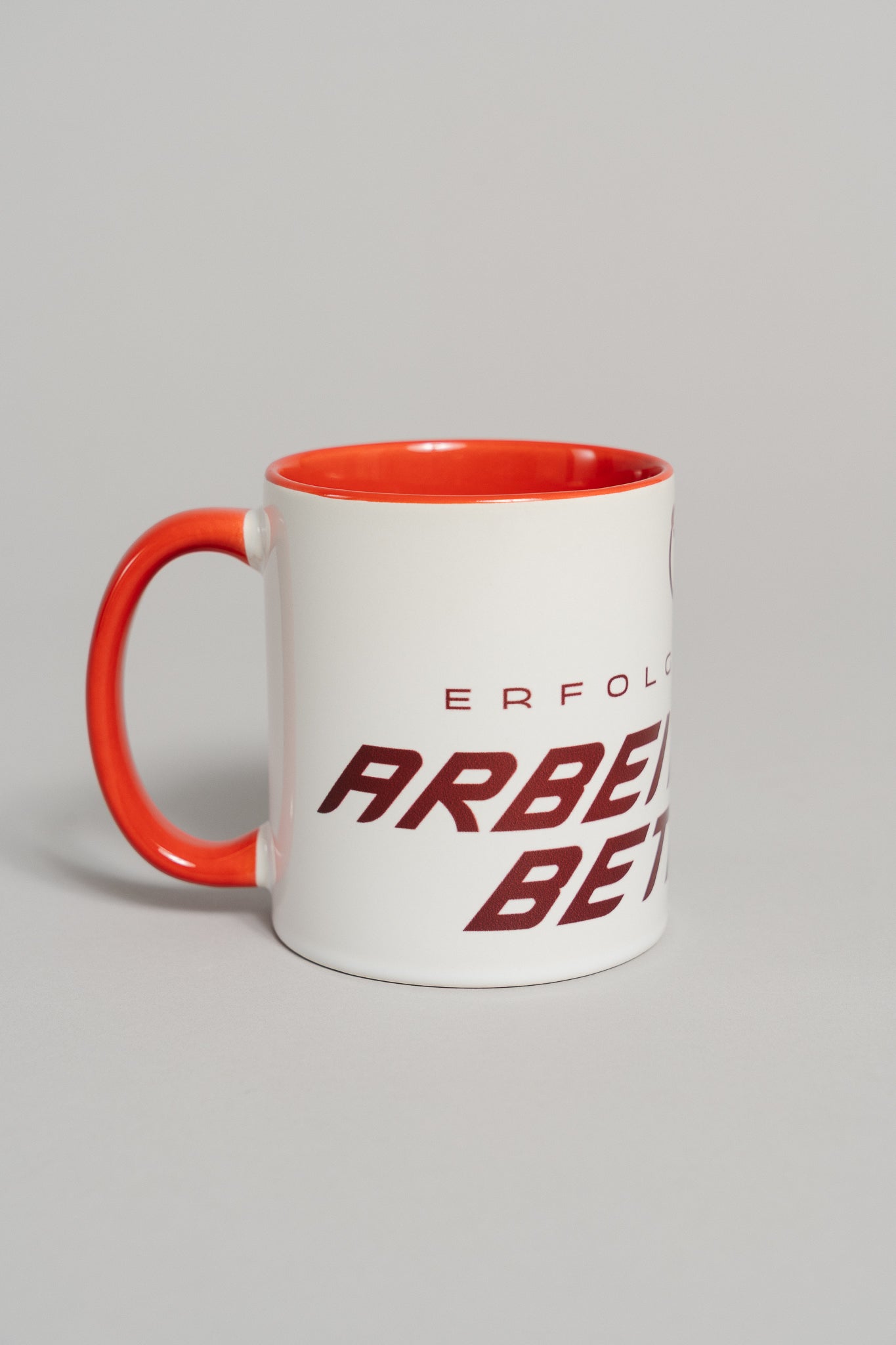 Weiße Keramiktasse mit rotem Henkel und rotem Innenbereich. Auf der Außenseite ist ein dunkler, schräg verlaufender Schriftzug zu sehen, der teilweise den Text „ERFOLGREICHER ARBEITSZEITBETRUG“ zeigt. Die Tasse steht auf einer neutral grauen Fläche und ist leicht seitlich gedreht fotografiert.