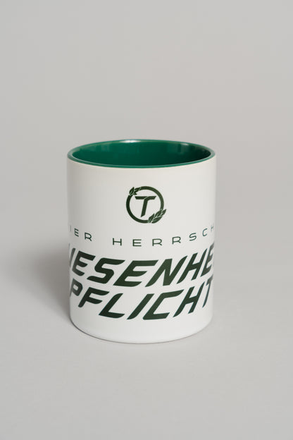 Weiße Keramiktasse mit dunkelgrünem Innenbereich. Auf der Vorderseite ist ein großer, schräg verlaufender Schriftzug in dunklem Grün zu sehen, der teilweise den Text „HIER HERRSCHT ANWESENHEITSPFLICHT“ zeigt. Darüber befindet sich ein rundes Logo mit dem Buchstaben „T“ und kleinen Blattelementen. Die Tasse steht auf einer neutralen grauen Fläche und wird frontal gezeigt.