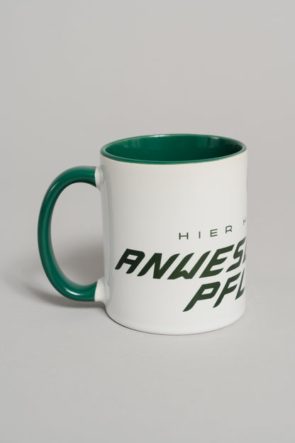 Weiße Keramiktasse mit grünem Henkel und grünem Innenbereich. Auf der Außenseite ist ein dunkler, schräg verlaufender Schriftzug zu sehen, der teilweise den Text „HIER HERRSCHT ANWESENHEITSPFLICHT“ zeigt. Die Tasse steht auf einer neutralen grauen Fläche und ist leicht seitlich gedreht, sodass Henkel und Schriftzug sichtbar sind.