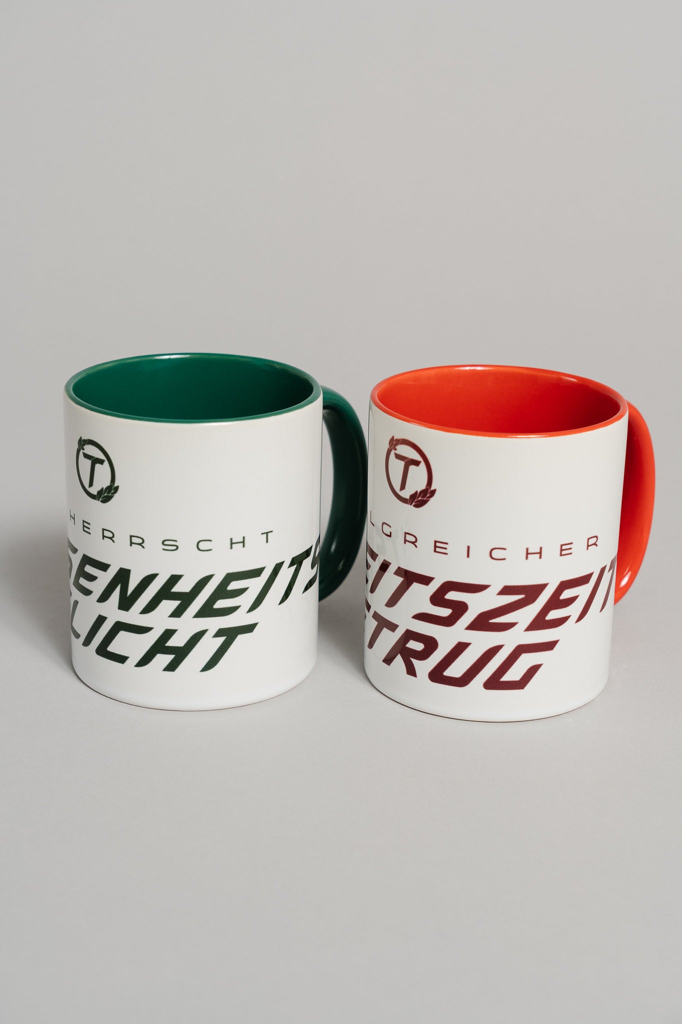 Zwei weiße Keramiktassen stehen nebeneinander auf grauer Fläche. Die rechte Tasse mit rotem Henkel und rotem Innenbereich trägt den dunkelroten Schriftzug „ERFOLGREICHER ARBEITSZEITBETRUG“ sowie ein rundes Logo mit dem Buchstaben „T“. Sie steht leicht im Vordergrund und ist der Hauptfokus des Bildes.