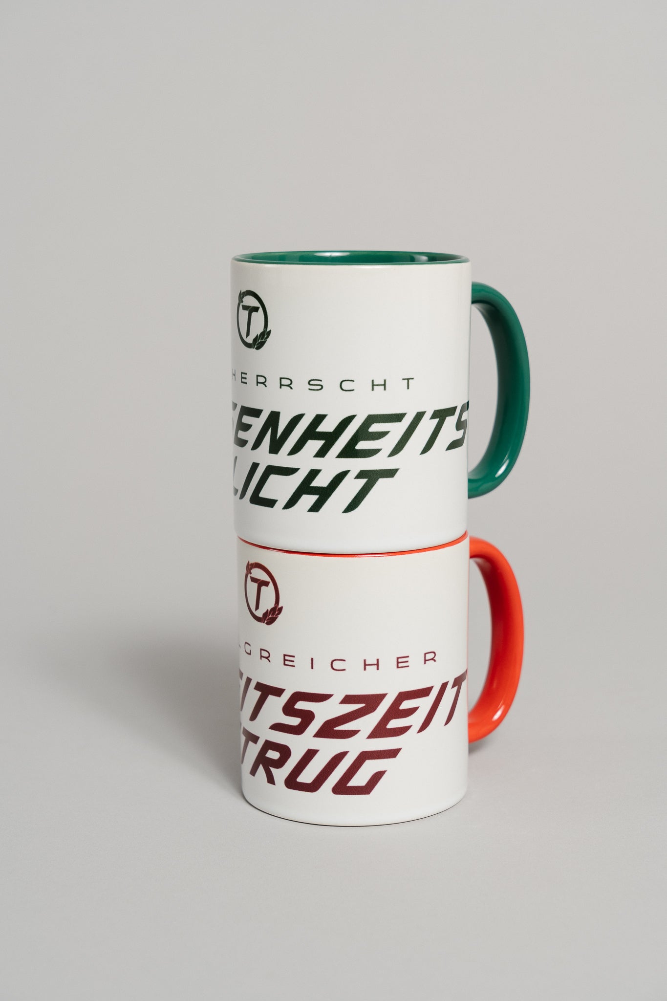 Zwei weiße Keramiktassen sind übereinandergestapelt. Die untere Tasse mit rotem Henkel und rotem Innenbereich steht im Fokus und trägt den dunkelroten Schriftzug „ERFOLGREICHER ARBEITSZEITBETRUG“ mit rundem „T“-Logo darüber. Darüber steht eine zweite Tasse mit grünem Henkel und grünem Innenbereich, die nur teilweise sichtbar ist.