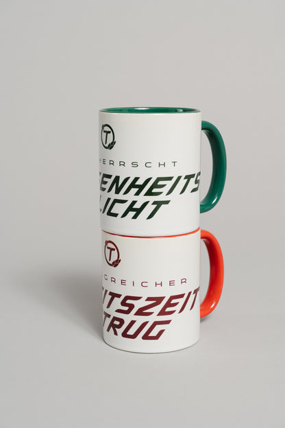 Zwei weiße Keramiktassen sind übereinandergestapelt. Die untere Tasse mit rotem Henkel und rotem Innenbereich steht im Fokus und trägt den dunkelroten Schriftzug „ERFOLGREICHER ARBEITSZEITBETRUG“ mit rundem „T“-Logo darüber. Darüber steht eine zweite Tasse mit grünem Henkel und grünem Innenbereich, die nur teilweise sichtbar ist.