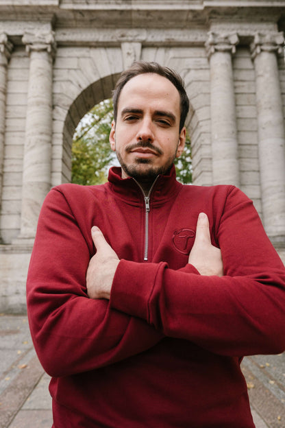 Ein burgunderrotes Sweatshirt mit kurzem Reißverschluss und hochgestelltem Kragen. Auf der linken Brust ist das ton-in-ton gestickte Logo mit dem Buchstaben „T“ im Lorbeerkranz zu sehen. Der Stoff wirkt weich und strukturiert. Die Person trägt das Sweatshirt mit verschränkten Armen und ernstem Blick, aufgenommen vor einem steinernen Torbogen im Freien.
