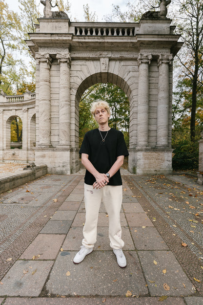 Schwarzes T-Shirt aus weicher Baumwolle mit gesticktem Ton-in-Ton-Logo auf der Brust. Das Design zeigt ein stilisiertes „T“ im Kreis mit Lorbeerzweigen. Das Shirt hat einen lockeren Schnitt und vermittelt einen cleanen, modernen Streetwear-Look, kombiniert mit heller Hose und sportiven Sneakern.