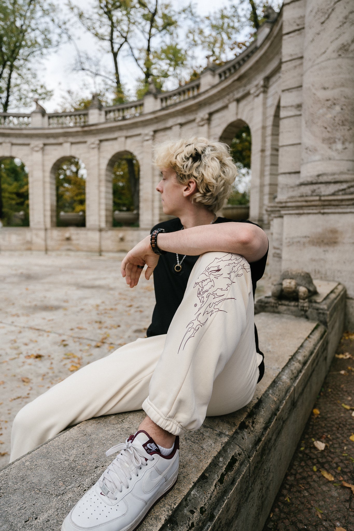 Beigefarbene Jogginghose mit braunrotem Linienprint auf dem rechten Bein, der ein stilisiertes Wolfsgesicht mit flammenartigen Konturen zeigt. Die Hose besitzt elastische Bündchen an den Knöcheln und wird im Sitzen getragen, wodurch das Motiv gut zur Geltung kommt.