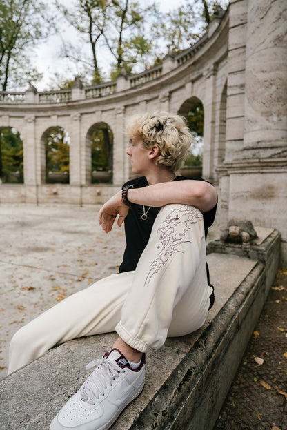 Beigefarbene Jogginghose mit braunrotem Linienprint auf dem rechten Bein, der ein stilisiertes Wolfsgesicht mit flammenartigen Konturen zeigt. Die Hose besitzt elastische Bündchen an den Knöcheln und wird im Sitzen getragen, wodurch das Motiv gut zur Geltung kommt.