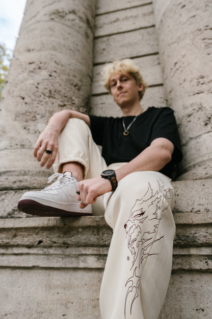 Beigefarbene Jogginghose aus Sweatstoff mit auffälligem, braunrotem Printmotiv auf dem rechten Bein. Der Druck zeigt ein stilisiertes, flammenförmiges Wolfsgesicht in dynamischer Linienführung. Die Hose hat elastische Bündchen an den Knöcheln und wird im Sitzen getragen, wodurch das Motiv gut sichtbar ist.